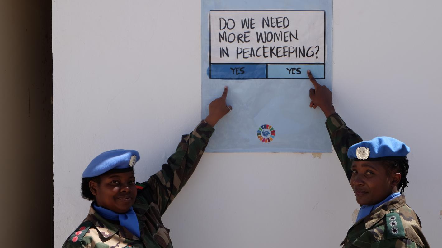 Two women peacekeepers in military uniform point at a sign that poses the question "do we need more women in peacekeeping" and gives "yes" or "yes" as possible responses. 
