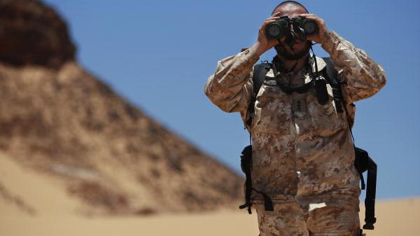 A UN peacekeeper in sand-coloured military uniform stands in the desert looking through binoculars
