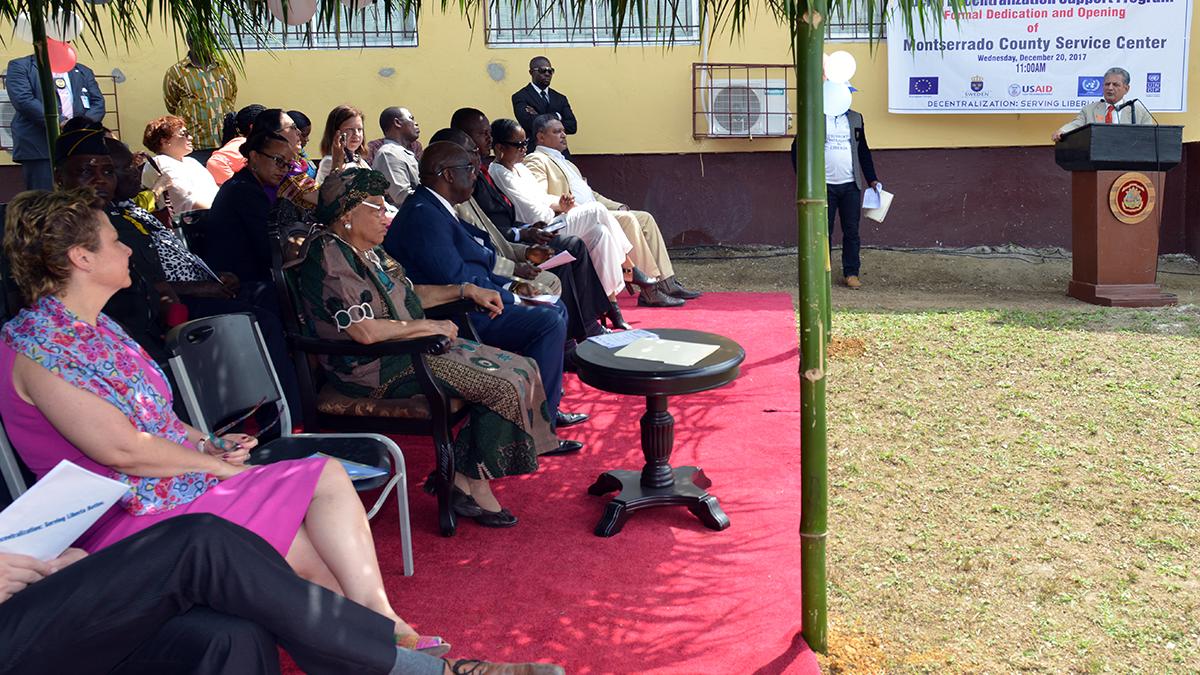 A group of officials sit outside County Service Centre, looking on as a man speaks at a podium