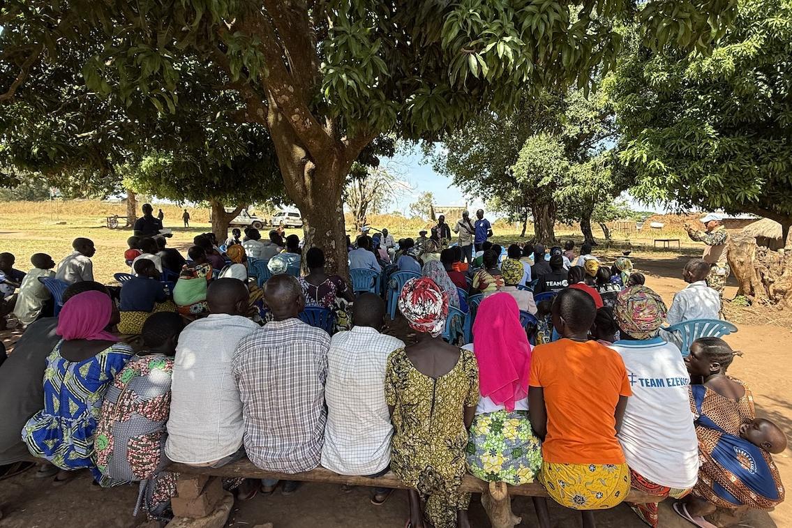 A group of people in brightly coloured clothing seated on wooden benches under a large tress are facing a person giving a presentation. 