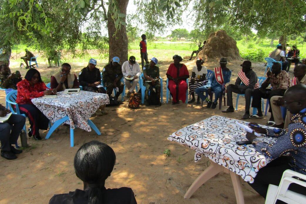 While patrolling in Panyijiar, Unity state, to help protect civilians and reduce tensions in the aftermath of air strikes, UNMISS peacekeepers hear from community members, who believe that dialogue is the only route towards a permanent peace in South Sudan. Group of people seated in a circle outdoors under a large tree, with two tables covered in patterned cloth at the center. Some participants wear blue caps, and traditional huts are visible in the background.