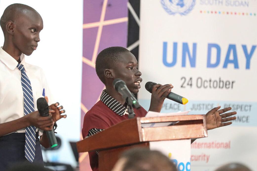 Two individuals speaking at a podium with microphones during a UN Day event, with a banner showing ‘UN Day 24 October’ in the background.