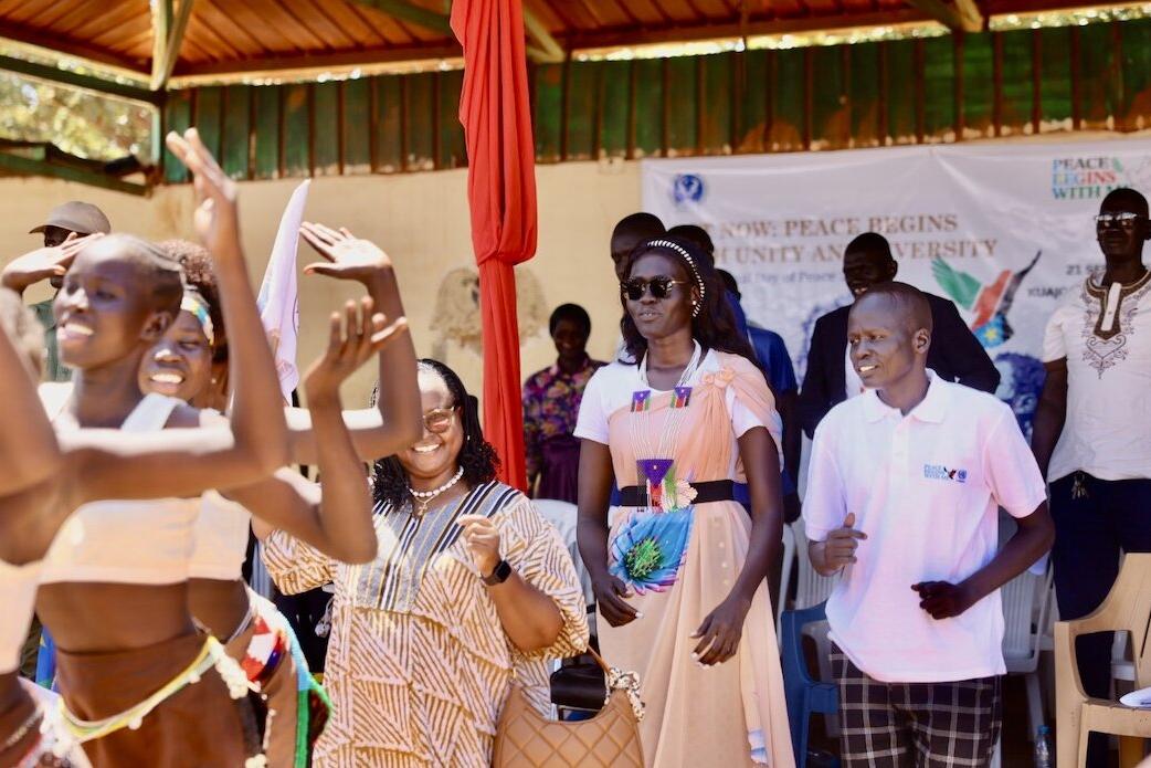 The crowd gathered at Kuajok's Freedom Square had a good time, embracing their Peace Day togetherness. People standing and dancing under a covered outdoor structure during an event, with a banner in the background displaying the words ‘Peace Begins’ and other text.